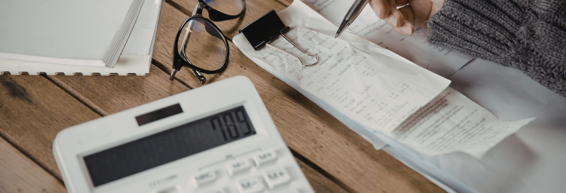 Man sieht einen Taschenrechner, Belege und eine Frauenhand, die Notizen macht.